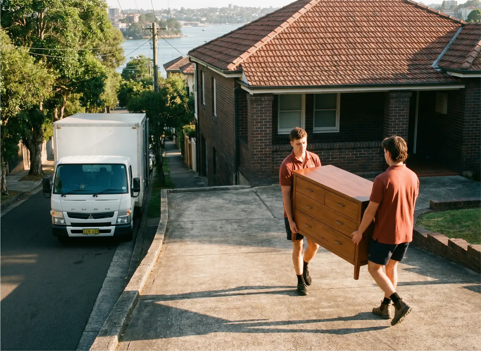 Two removalists carrying a chest of drawers up a steep driveway to a brick house